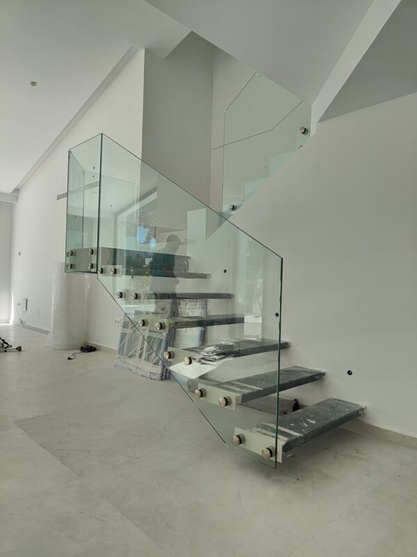 Escalera interior suspendida fabricada mediante carpintería metálica, con peldaños anclados a paneles de vidrio.